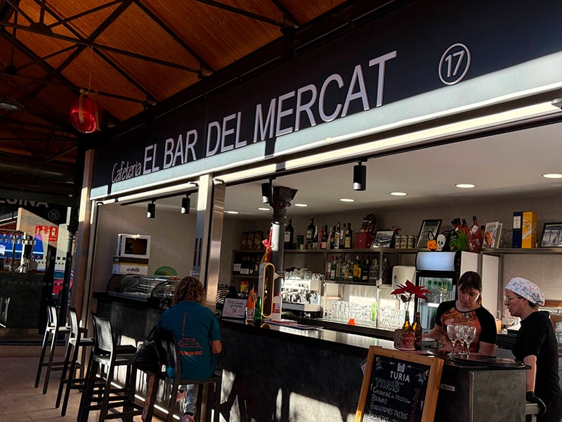 Барная стойка El Bar del Mercat внутри рынка: вывеска, напитки на полках и посетители у стойки (Кабаньяль, Валенсия).