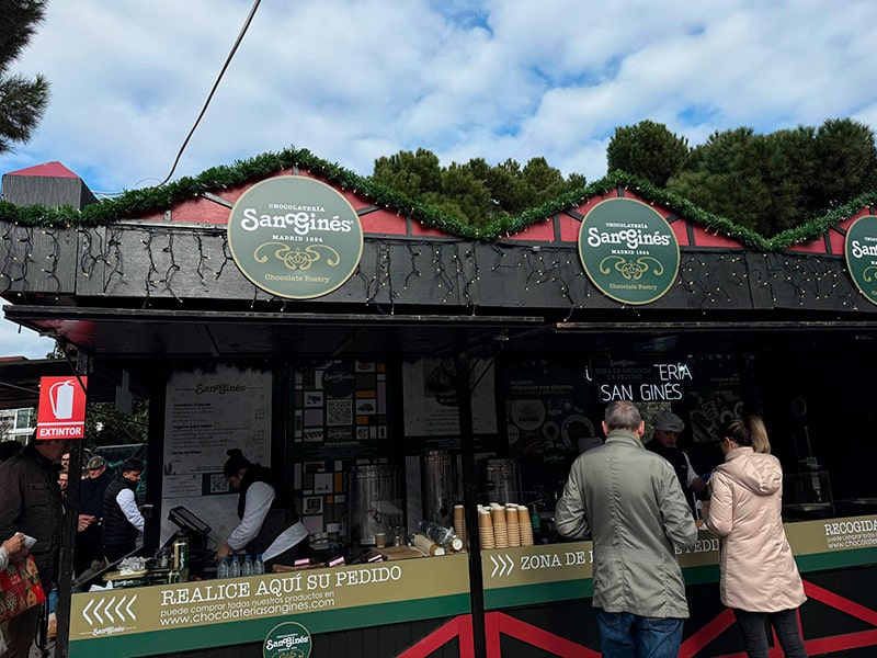 Рождественний киоск Chocolater&iacute;a San Gin&eacute;s на Plaza de Col&oacute;n в Мадриде: посетители заказывают чуррос и горячий шоколад у стойки.
