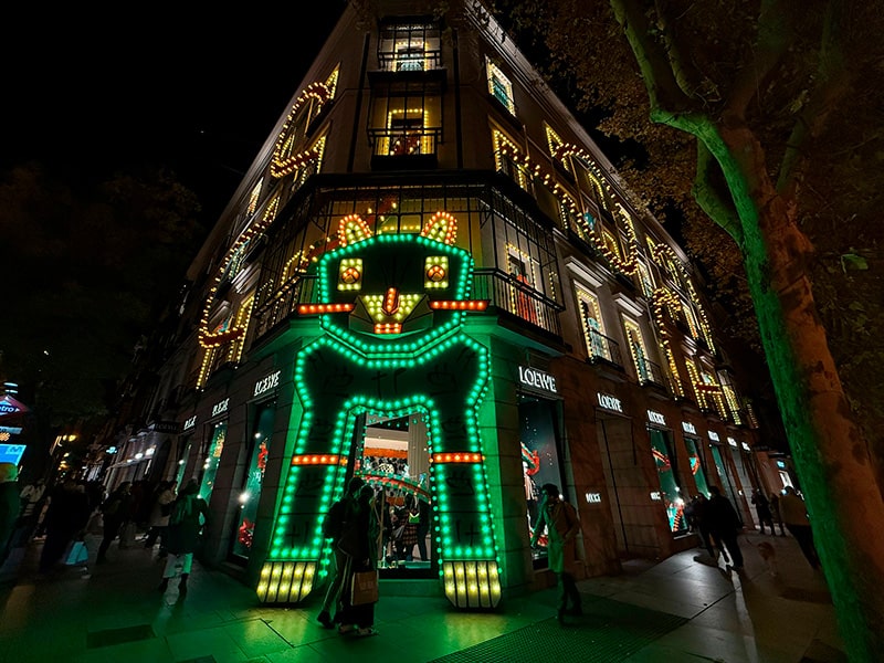 Рождественская иллюминация на фасаде магазина LOEWE на Gran V&iacute;a в Мадриде, светящийся портал в виде зеленого кота у входа, огни по всему зданию и прохожие вечером