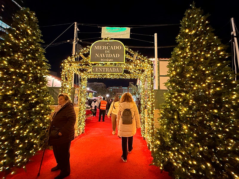 Вход на Mercado de Navidad El Corte Ingl&eacute;s в Мадриде: арка с гирляндами, красная ковровая дорожка и люди, входящие на рождественский рынок