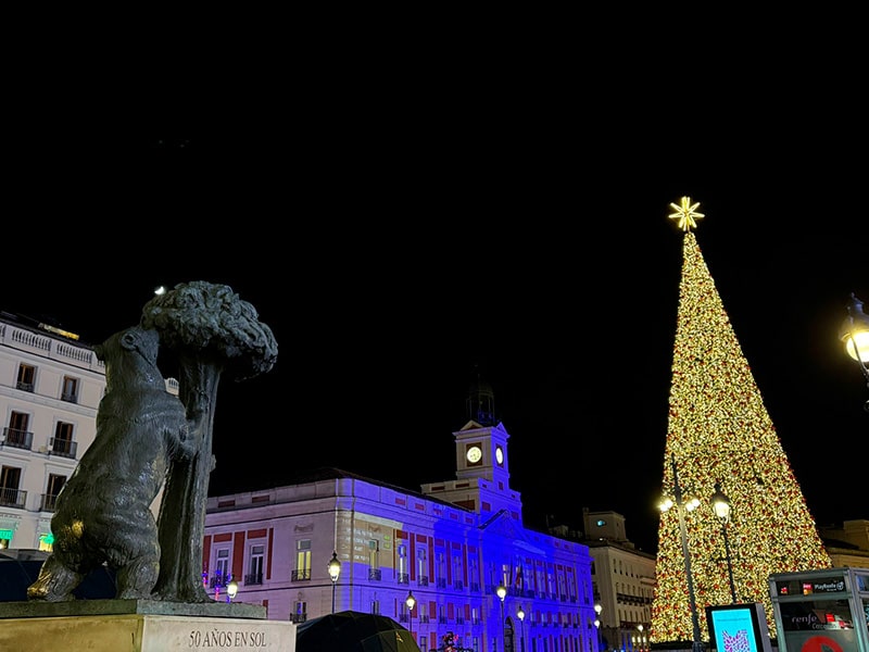 Рождественская елка на Puerta del Sol в Мадриде, справа высокая сверкающая елка со звездой, слева статуя медведя и мадроньо, на заднем плане подсвеченное здание Real Casa de Correos