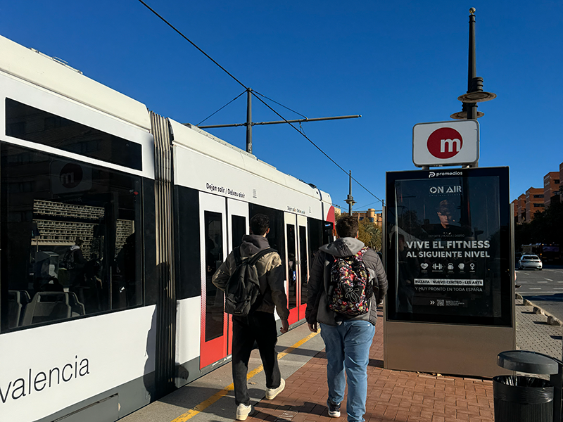 Остановка Metrovalencia в районе Algir&oacute;s (Валенсия): двое людей с рюкзаками идут вдоль платформы рядом с бело-красным поездом, рядом знак метро и рекламный стенд.