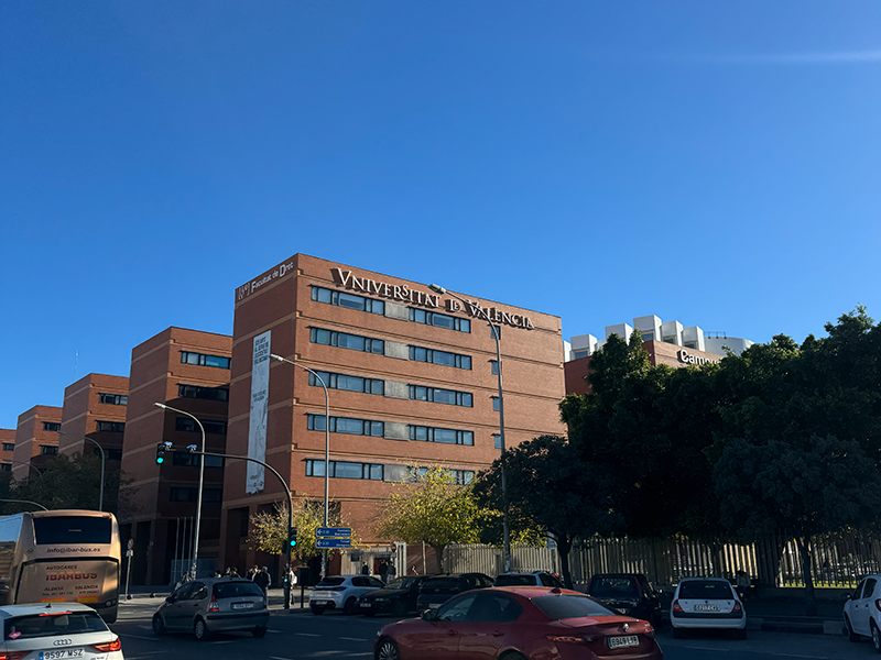 Здание Universitat de Val&egrave;ncia в районе Algir&oacute;s, Валенсия: кирпичный корпус университета на фоне ясного голубого неба, перед ним городская улица с автомобилями.