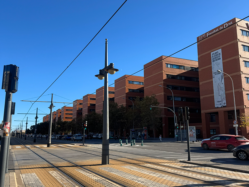 Трамвайные пути и остановка рядом с кампусом Universitat de Val&egrave;ncia в районе Algir&oacute;s: кирпичные корпуса факультетов вдоль проспекта, ясное небо и городское движение.